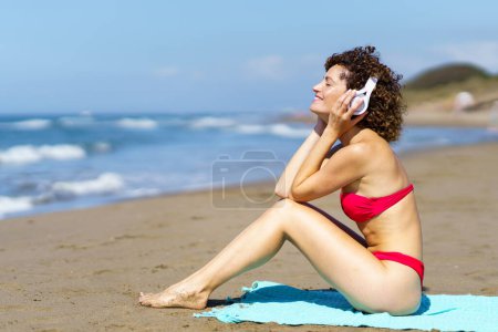 Foto de Vista lateral de la alegre hembra en bikini rosa y con los ojos rizados cerrando el pelo mientras está sentada en una manta en la playa de arena, y escuchando música con auriculares - Imagen libre de derechos