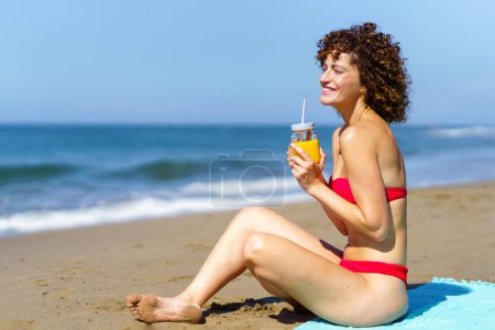 Téléchargez les photos : Vue latérale de la jeune femme aux cheveux bouclés et en bikini assis sur une plage de sable avec un verre de jus d'orange et regardant loin - en image libre de droit