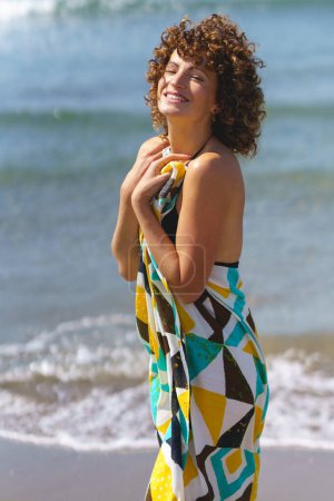 Téléchargez les photos : Vue latérale de jeune femme sensuelle heureuse avec des cheveux bouclés en robe colorée debout sur le bord de la mer et regardant la caméra - en image libre de droit