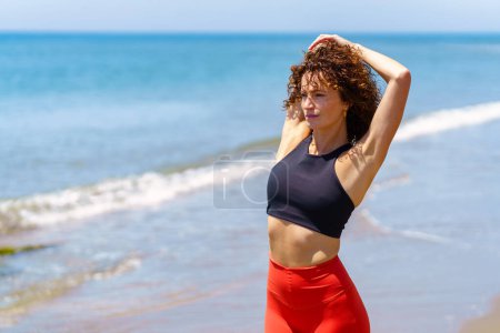 Téléchargez les photos : Jeune athlète réfléchie en vêtements de sport debout près de la mer et étirant le bras derrière le dos tout en regardant loin - en image libre de droit