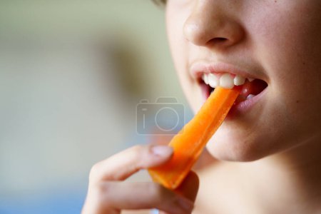 Téléchargez les photos : Gros plan de culture anonyme jeune fille mangeant des tranches de carotte biologique fraîche à la maison - en image libre de droit