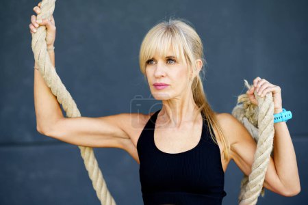 Photo for Young fit female wearing sportswear holding rope against gray background in fitness studio looking away thoughtfully while exercising at modern gym - Royalty Free Image