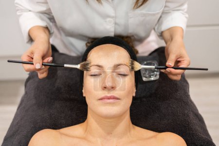 Photo for From above crop beauty specialist applying liquid cosmetic with brushes on face of female customer in modern salon during skin care treatment - Royalty Free Image