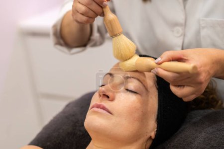 Téléchargez les photos : Crop cosmétique professionnel massant le front du client féminin avec des brosses pendant le traitement de la peau dans le centre de beauté moderne - en image libre de droit