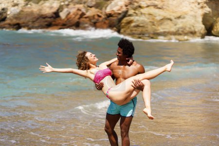Téléchargez les photos : Vue latérale d'une jeune femme joyeuse étirant le bras vers l'arrière, allongée dans les mains d'un homme afro-américain debout et se regardant près de l'eau de mer pendant la journée - en image libre de droit