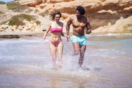 Photo for Full body of multiracial couple in swimwear running in splashing water and having fun while looking away and enjoying free time near seaside in summertime - Royalty Free Image