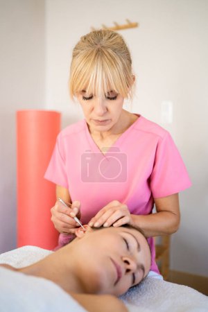 Téléchargez les photos : Chiropracteur esthéticienne professionnelle faisant massage de l'oreille profonde de la jeune cliente avec un stylo à sonde auditive pendant le traitement de la peau dans un salon de beauté moderne - en image libre de droit