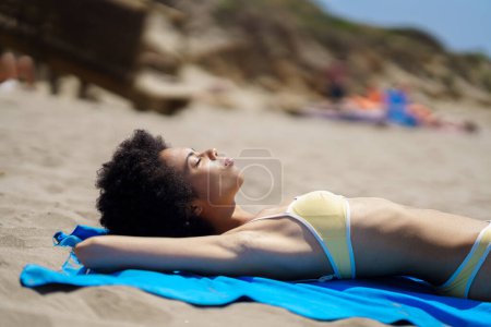 Téléchargez les photos : Vue latérale d'une jeune voyageuse afro-américaine calme aux cheveux bouclés, en maillot de bain élégant profitant d'une journée d'été ensoleillée allongée sur une serviette bleue sur une plage de sable avec les yeux fermés - en image libre de droit
