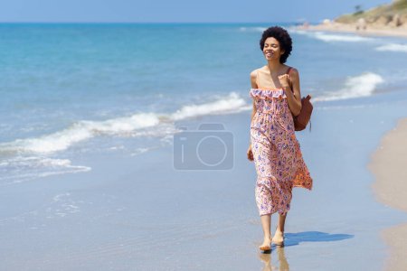 Foto de Cuerpo completo de joven mujer afroamericana en vestido colorido sonriendo mientras pasa las vacaciones de verano en la playa de arena cerca del mar ondulante y mirando hacia otro lado - Imagen libre de derechos