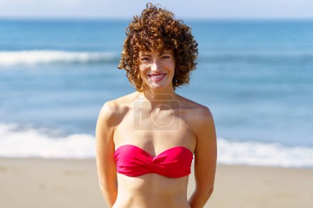 Téléchargez les photos : Portrait de jeune femme heureuse en bikini rose avec les cheveux bouclés souriant et regardant la caméra tout en se tenant sur la plage près de la mer agitant - en image libre de droit