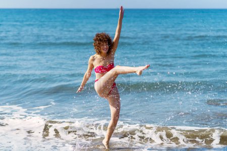 Téléchargez les photos : Corps complet de femme ludique en bikini donnant un coup de pied à l'eau de mer et éclaboussant sur la plage tout en profitant d'une journée d'été ensoleillée - en image libre de droit