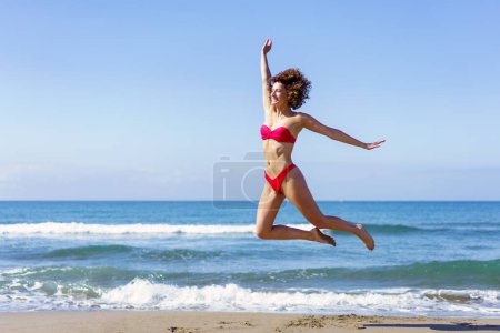 Téléchargez les photos : Vue latérale complète du corps de la jeune femme excitée pieds nus, en bikini avec les bras tendus bondissant avec les jambes dans l'air tout en regardant loin dans la lumière du jour contre l'eau de mer ondulante mousseuse - en image libre de droit