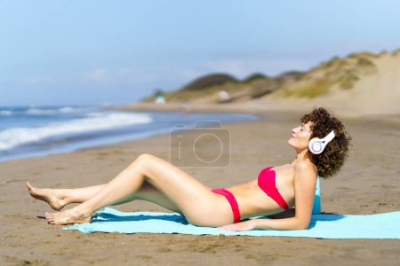 Téléchargez les photos : Vue latérale de la femelle mince en bikini rouge se prélasser sur la plage et écouter de la musique dans les écouteurs sous un soleil éclatant - en image libre de droit