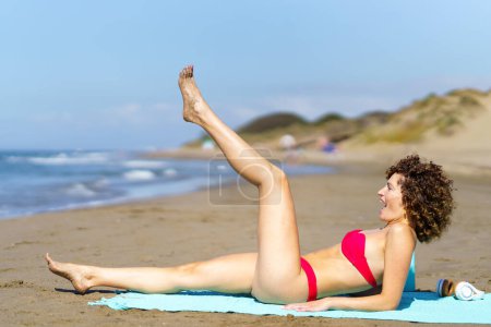 Foto de Vista lateral de una mujer joven con el pelo rizado en bikini rosa levantando la pierna mientras se divierten en la costa arenosa - Imagen libre de derechos