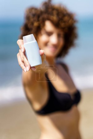 Téléchargez les photos : Foyer doux de la femme en bikini noir debout sur la plage près de la mer et démontrant tube vide de crème solaire - en image libre de droit