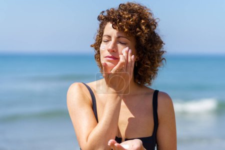 Téléchargez les photos : Jeune femme aux cheveux bouclés en bikini noir appliquant de la crème solaire sur les joues tout en se tenant près de la mer lors d'une journée d'été ensoleillée protégeant la peau des coups de soleil - en image libre de droit