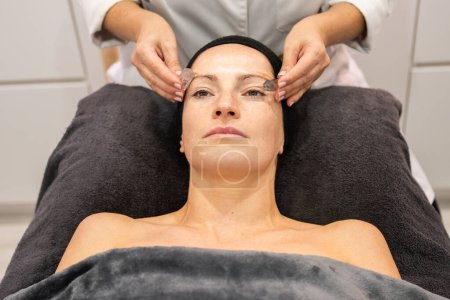 Photo for From above of woman lying face up on black pillow receiving facial skin treatment from crop masseuse in beauty center in daytime - Royalty Free Image