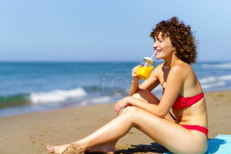 Téléchargez les photos : Vue latérale de la jeune rousse en bikini assise sur la côte et buvant du jus d'orange au soleil - en image libre de droit