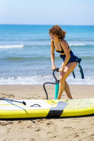 Téléchargez les photos : Jeune femme souriante aux cheveux bouclés au gingembre, en maillot de bain debout près du paddleboard et pompant de l'air avec pompe à main tout en regardant loin contre une mer agitée floue et un ciel bleu - en image libre de droit