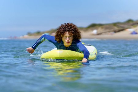 Téléchargez les photos : Jeune surfeuse concentrée en combinaison avec les cheveux bouclés couchés sur la planche SUP tout en pagayant avec les mains flottant sur l'océan pendant les vacances d'été - en image libre de droit