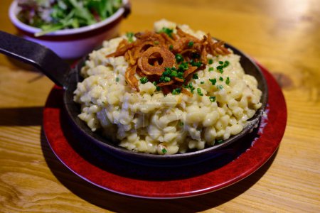 Photo for Pongauer Kasnocken Cheese Spaetzle Pasta in an Iron Pan with Fried Crispy Onion and Green Salad - Royalty Free Image