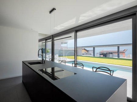 Photo for Black modern kitchen with sink and stools. There are bright windows where lots of light comes in and you can see the pool and garden. Nobody inside - Royalty Free Image