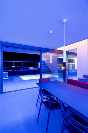 Photo for Liiving room with table and a red sofa lit by blue LEDs and lamps attached to the ceiling. Large window overlooking the lighted pool and garden. Nobody inside - Royalty Free Image