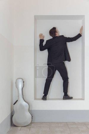 Téléchargez les photos : Un jeune homme en costume élégant se tient debout dans un cadre contre le mur. À côté de lui se trouve son étui à guitare. Isolé sur fond blanc - en image libre de droit