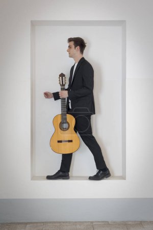 Photo for A young boy in an elegant suit is standing in a niche in the wall, trying to get out. He is holding his guitar with one hand. Isolated on white background - Royalty Free Image
