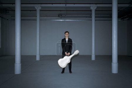 Photo for A boy is standing with his guitar case, which he holds with both hands. Interior of a renovated former factory, large columns - Royalty Free Image