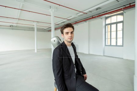 Photo for A young boy sits on a stool, carrying his guitar case over his shoulder. Interior of a renovated former factory. - Royalty Free Image