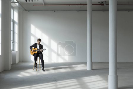 Téléchargez les photos : Portrait d'un jeune garçon jouant de la guitare. Il porte un costume noir. Intérieur d'une ancienne usine rénovée. - en image libre de droit