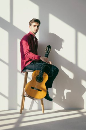 Photo for A young boy is sitting on a stool, holding his guitar with one hand. He is wearing a white shirt and a red shirt. Interior of a renovated former factory. - Royalty Free Image