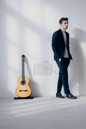 Photo for A standing boy is walking away from his guitar with his hands in his pockets. He is wearing a black suit. Interior of a renovated former factory. - Royalty Free Image