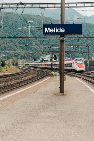 Foto de Estación Melide, señal azul con el nombre de la estación. Viene el tren de alta velocidad. En el fondo están las montañas con los árboles verdes. - Imagen libre de derechos
