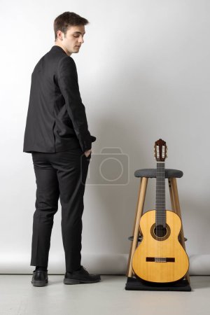 Téléchargez les photos : Un jeune garçon en costume noir regarde sa guitare appuyée sur un tabouret à côté de lui. Fond blanc - en image libre de droit