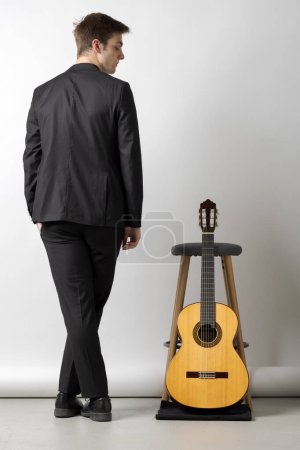Photo for Young boy from behind in a black suit looks at his guitar leaning on a stool next to him. White background - Royalty Free Image