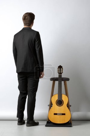 Téléchargez les photos : Jeune garçon par derrière en costume noir, appuyé sur un tabouret avec sa guitare à côté de lui. Fond blanc - en image libre de droit