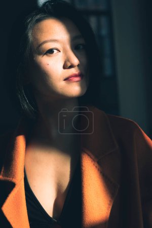 Téléchargez les photos : Portrait d'une jeune femme chinoise réfléchie portant un beau manteau orange. Contraste fort entre lumière et ombre - en image libre de droit