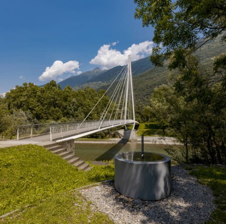 Photo for Modern pedestrian bridge in the middle of the mountains of Italian Switzerland, which allows you to cross the Ticino river. Beautiful sunny summer day. There is a modern fountain in the foreground - Royalty Free Image