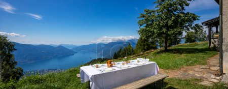 Téléchargez les photos : Table imn extérieur avec une grande nappe blanche et une vue sur le lac Majeur. Une vue magnifique, vous pouvez même voir les îles Brissago - en image libre de droit