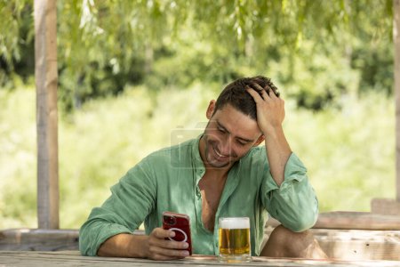 Photo for Young, handsome guy in the middle of summer sitting at a table with a beer in nature while looking at his phone smiling. - Royalty Free Image