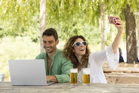 Téléchargez les photos : Jeune couple adulte en plein air au milieu de l'été à Lugano, il regarde son ordinateur sourire et elle prend un selfie, deux belles bières froides sont vues sur la table. Ils travaillent et s'amusent. Ils sont très heureux. - en image libre de droit