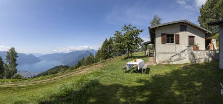 Téléchargez les photos : Table avec assiettes et couverts au milieu d'une prairie, vue magique sur le lac Majeur. Une maison de vacances d'été suisse. Journée ensoleillée au milieu de l'été. - en image libre de droit