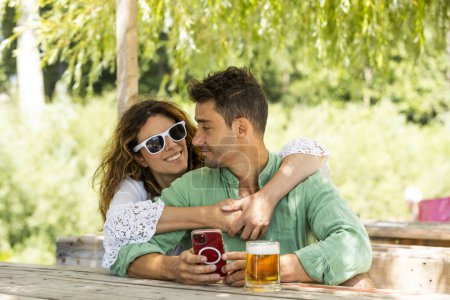 Téléchargez les photos : Jeune couple hétérosexuel assis à une table extérieure au milieu de l'été tout en regardant un téléphone mobile. Devant eux, une bonne bière fraîche. Elle porte des lunettes de soleil. - en image libre de droit