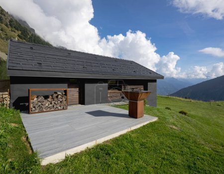 Photo for A very unusual Swiss mountain chalet because it is black and made of wood. Surrounded by a beautiful green summer meadow. The sky is blue and it looks like a quiet and very peaceful place. A very modern grill for cooking meat outside. - Royalty Free Image