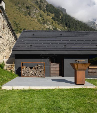 Téléchargez les photos : Un chalet de montagne suisse très insolite car il est noir et en bois. Entouré d'une belle prairie verte d'été. Le ciel est bleu et il ressemble à un endroit calme et très paisible. Un grill très moderne pour cuisiner la viande à l'extérieur. - en image libre de droit