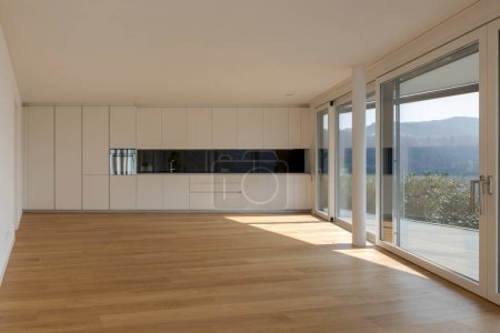 Photo for Modern living room with open space and a black and white designed kitchen situated at the end. Large windows allow plenty of sunlight to enter. The room features a parquet floor. Nobody inside - Royalty Free Image