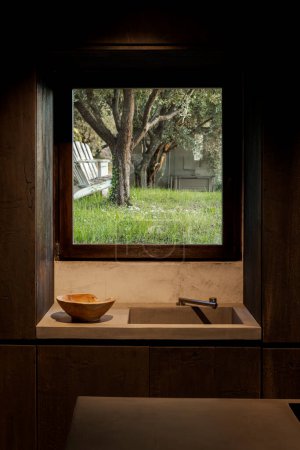 Téléchargez les photos : Détail de cuisine contemporaine en bois sombre avec un évier à côté d'un bol. La fenêtre donnant sur le jardin offre une vue sur les oliviers. Personne n'est présent.. - en image libre de droit