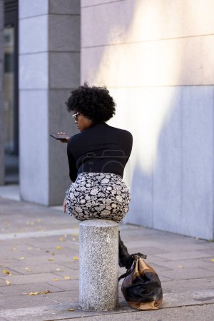 Téléchargez les photos : Femme afro très sexy assis sur un pieu en ville est au téléphone regardant vers le bas. Sur le sol est son sac à main. Scène urbaine. - en image libre de droit